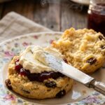 A close-up of a warm, split fruit scone being spread with thick clotted cream and strawberry jam, with steam rising from the crumbly center.