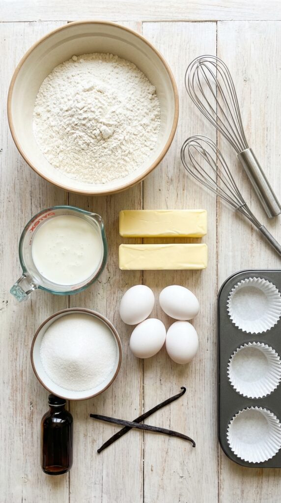 An overhead flat lay of baking ingredients including flour, buttermilk, butter, eggs, sugar, and vanilla beans on a wooden table.