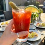 A close-up of a hand holding a sweating bloody mary glass with a pickle garnish in a brunch setting.