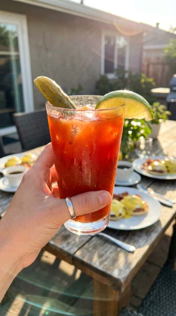 A close-up of a hand holding a sweating bloody mary glass with a pickle garnish in a brunch setting.