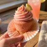 A close-up of a hand unwrapping a pink cupcake liner, revealing a moist pink cake crumb topped with pink frosting.