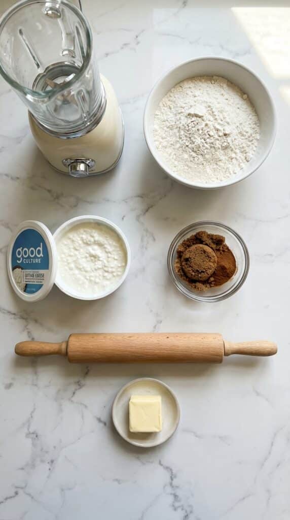 A flat lay showing cottage cheese, flour, brown sugar, cinnamon, butter, and a rolling pin on a marble surface.