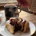 A close-up of a fork pulling apart a warm, gooey cinnamon roll covered in icing, with a coffee mug in the background.