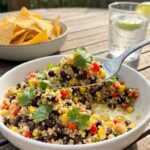 A close-up of a fork lifting a bite of colorful quinoa and black bean salad with tortilla chips in the background.