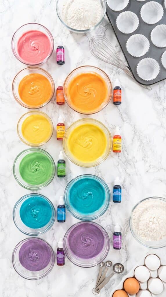 A flat lay showing six bowls of bright rainbow-colored cake batter, food coloring bottles, and baking ingredients on a marble board.