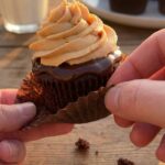 A close-up of a hand peeling the wrapper off a chocolate cupcake with tall peanut butter frosting and chocolate drizzle.