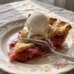 A slice of rhubarb pie topped with melting vanilla ice cream on a floral plate, with a fork cutting into the flaky crust.