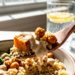 A close-up of a fork lifting a bite of quinoa, sweet potato, and a chickpea coated in tahini dressing from a bowl.