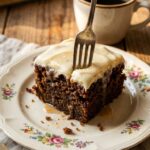 A close-up of a fork cutting into a moist square of glazed carrot cake with cream cheese frosting.