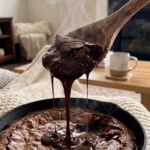 close-up of a spoon lifting a serving of brownie pudding, revealing thick, dripping chocolate sauce underneath the cake layer.