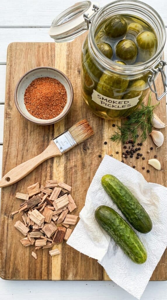 A flat lay showing a jar of pickles, BBQ rub, wood chips, and a paper towel on a wooden board.