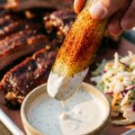 A close-up of a hand dipping a spiced smoked pickle spear into a bowl of ranch dressing.