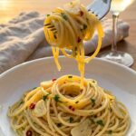 A close-up of a fork twirling glossy spaghetti with garlic and parsley, lifted from a white bowl.