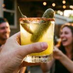 A hand holding a spicy pickle margarita with a tajin rim for a toast, with a blurred party background.