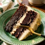 A close-up of a slice of dark chocolate cake with pale frosting on a green plate, with a gold fork taking a bite.