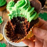 A close-up of a hand holding a chocolate cupcake with green frosting and gold sprinkles, with a bite taken out of it to show the moist crumb.