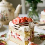 A close-up of a slice of strawberry cream cake on a floral plate with a fork taking a bite.