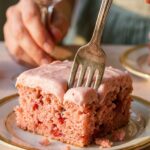 A close-up of a square slice of pink strawberry cake with a fork, and a glass of wine in the background.