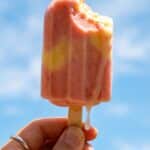 A close-up of a hand holding a bitten pink popsicle against a blue sky, showing a creamy texture.