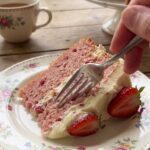 A close-up of a fork cutting into a moist slice of pink strawberry cake on a floral plate.