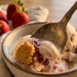 A close-up of a spoon lifting a bite of melting strawberry cheesecake ice cream with a crust chunk.