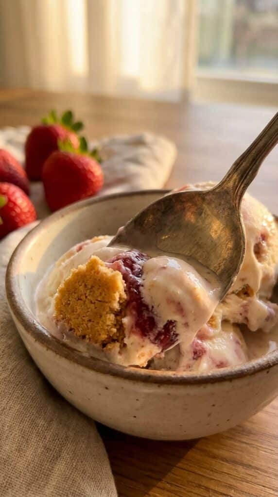 A close-up of a spoon lifting a bite of melting strawberry cheesecake ice cream with a crust chunk.