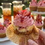 A close-up of a hand holding a vanilla cupcake topped with a tall swirl of pink strawberry frosting at an outdoor party.