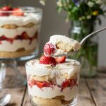 A close-up of a glass cup with a serving of strawberry trifle, with a spoon lifting a bite of cake and cream.