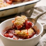 A close-up of a spoon lifting a bite of warm strawberry cobbler with steam rising