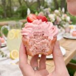 A close-up of a hand holding a bitten strawberry cupcake, showing the moist pink interior crumb.