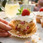 A close-up of a hand peeling the wrapper off a strawberry cupcake, revealing the fruit inside.