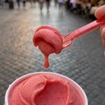 A close-up of a spoon lifting a bite of melting strawberry gelato from a cup.