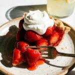 A side view of a slice of strawberry glaze pie showing whole berries suspended in red gel, served with whipped cream.