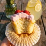 A close-up of a hand holding a bitten lemon cupcake with pink frosting, showing the moist yellow interior.