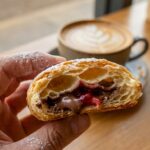 A close-up of a hand holding a bitten puff pastry tart, showing flaky layers and gooey chocolate filling.