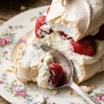 A close-up of a spoon breaking the crisp shell of a pavlova slice to reveal the soft center.