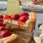 A close-up of a slice of strawberry pizza being lifted, showing the cookie crust and cream cheese layers.