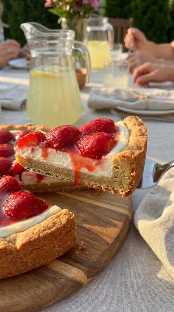 A close-up of a slice of strawberry pizza being lifted, showing the cookie crust and cream cheese layers.