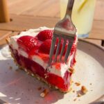 A close-up of a fork cutting into a slice of layered strawberry pretzel dessert, breaking the crust.