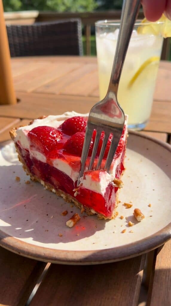 A close-up of a fork cutting into a slice of layered strawberry pretzel dessert, breaking the crust.