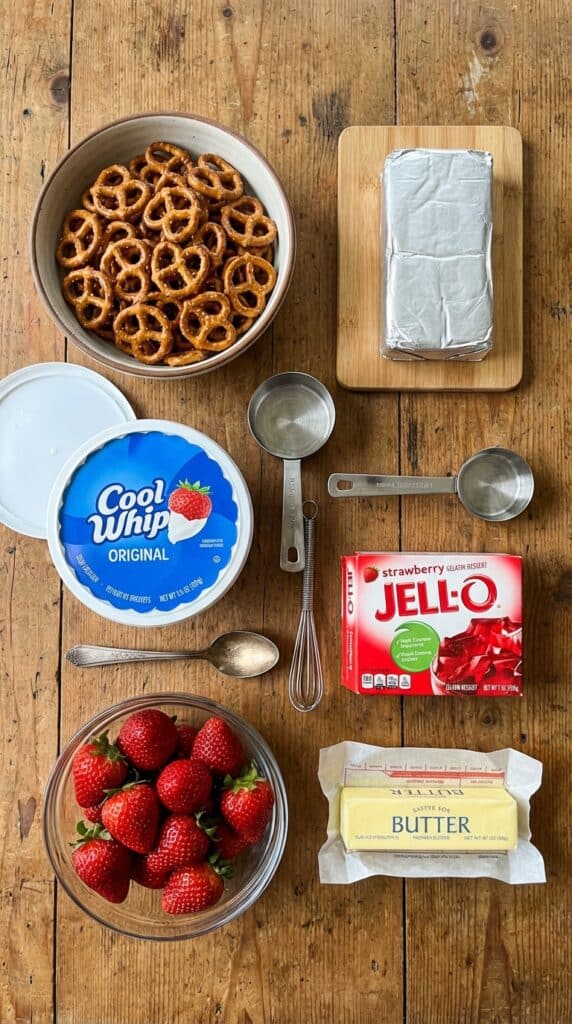 A flat lay showing pretzels, cream cheese, cool whip, jello box, and fresh strawberries on a wooden table.