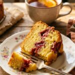 A close-up of a slice of strawberry rhubarb cake on a plate with a fork, showing the moist interior and fruit pockets.