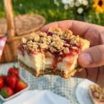 A close-up of a hand holding a cheesecake bar with a bite taken out, showing the creamy fruit filling.