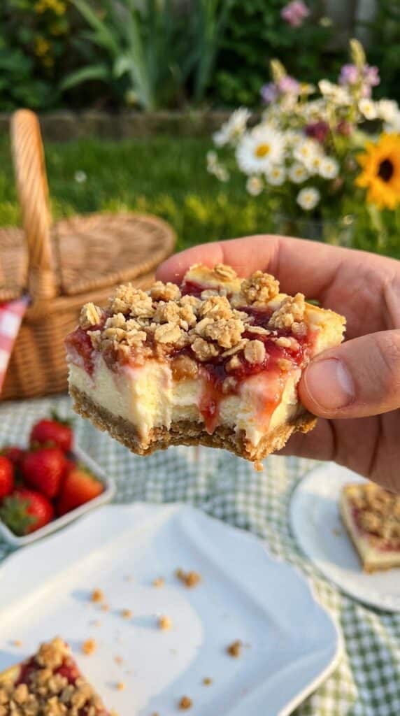 A close-up of a hand holding a cheesecake bar with a bite taken out, showing the creamy fruit filling.