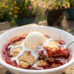 A close-up of a bowl of warm cobbler with melting vanilla ice cream and a spoon.