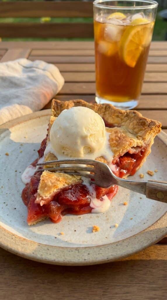 A close-up of a slice of strawberry rhubarb pie topped with melting vanilla ice cream.