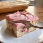A close-up of a square slice of strawberry cake on a plate, showing strawberry pieces inside the crumb.