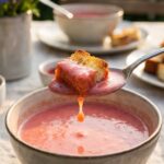 A close-up of a spoon lifting a piece of toasted cake and pink strawberry soup.