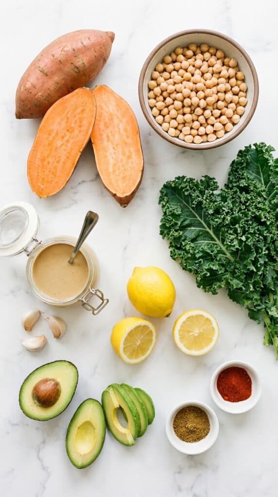 A flat lay showing raw sweet potatoes, chickpeas, kale, tahini, lemons, avocado, and spices on a white marble board.