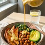A close-up of thick, creamy tahini dressing being poured from a small pitcher over a bowl of sweet potatoes, chickpeas, and avocado.